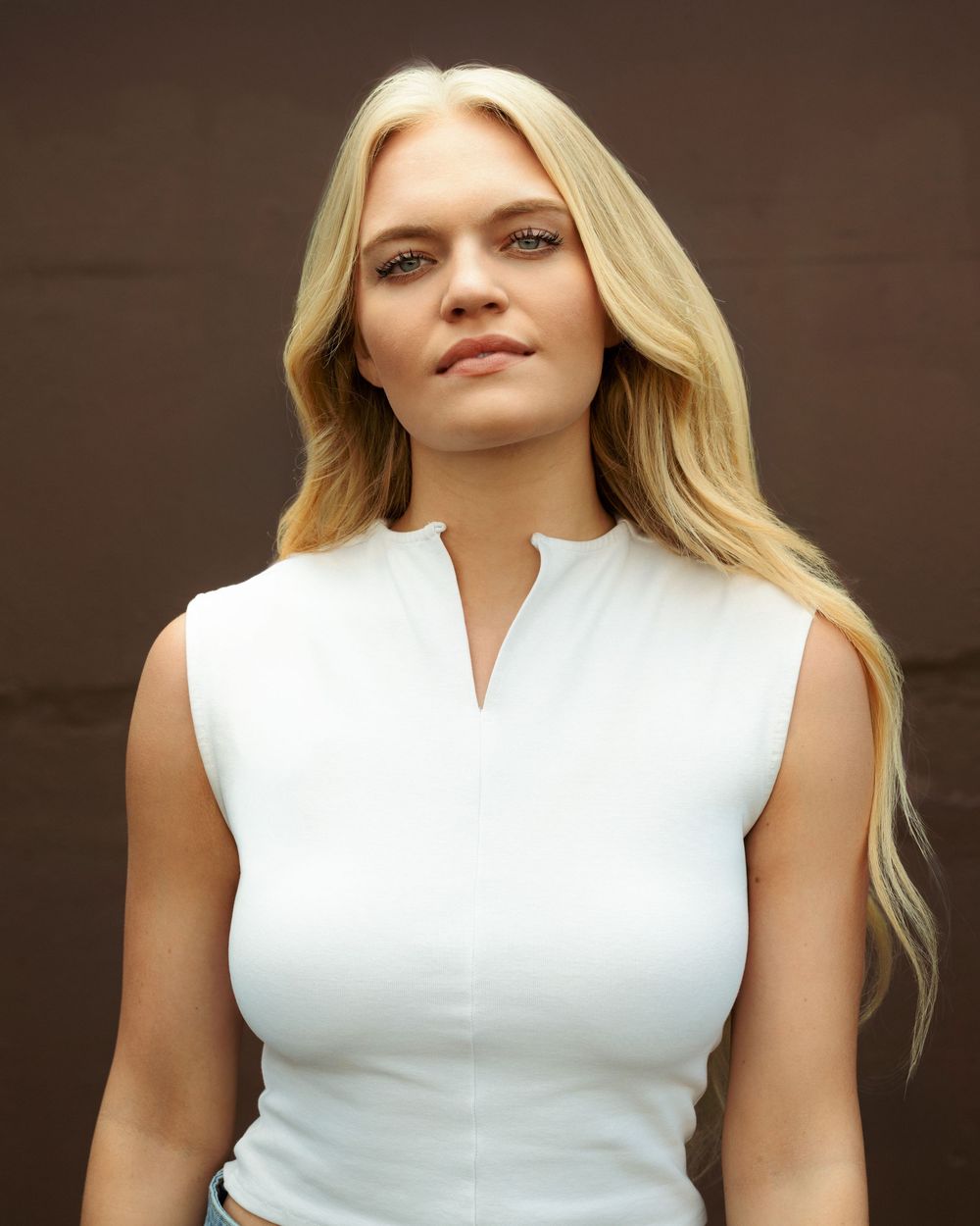 Young blonde woman in white sleeveless top posing confidently against a dark brown background.
