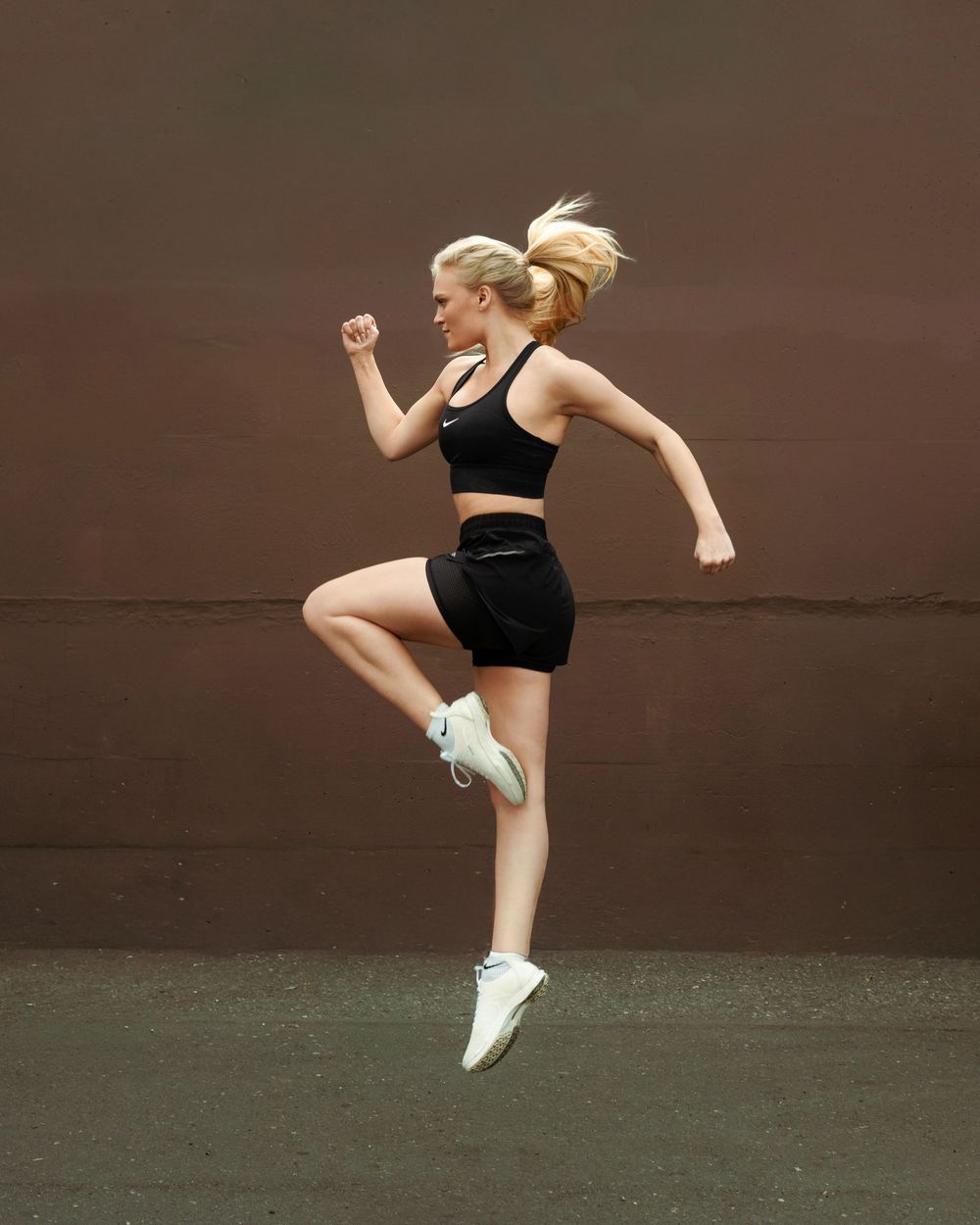 Blonde woman in black Nike sports bra and shorts jumping mid-air against a brown wall outdoors.
