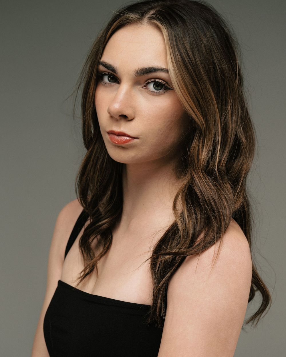 Young woman with long brown highlighted hair wearing a black strapless top, posing against a gray studio background.