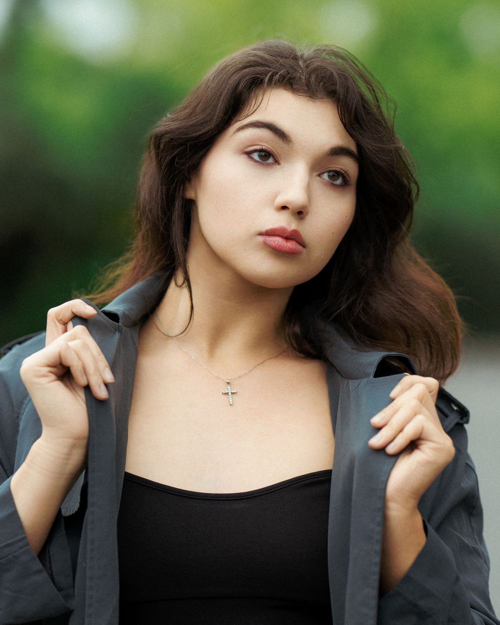 Young woman with dark hair wearing a black top, gray jacket, and cross necklace posing outdoors.