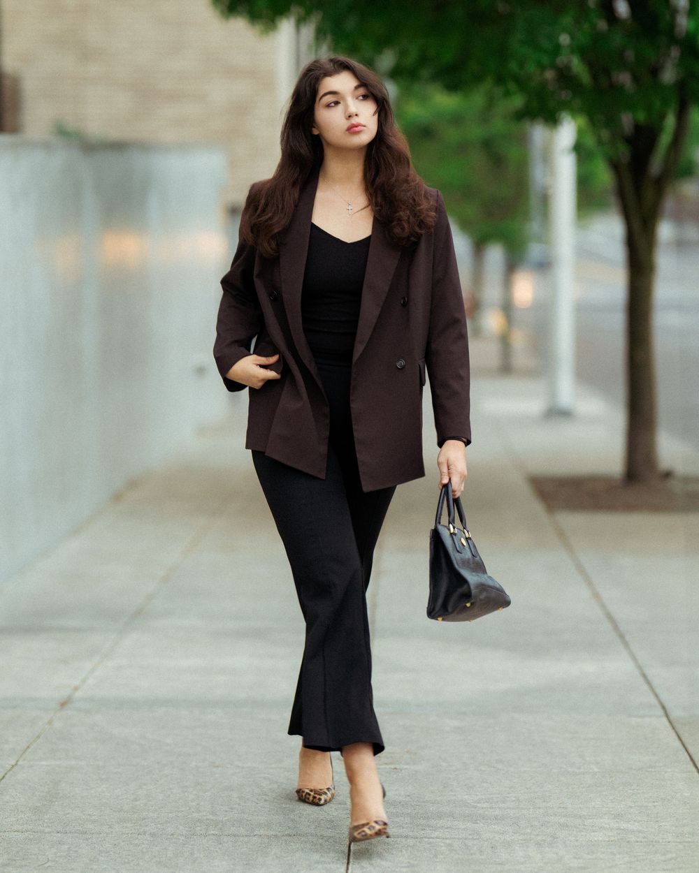 Woman in dark blazer, black top, trousers, and leopard heels walks confidently on a city sidewalk carrying a black bag.