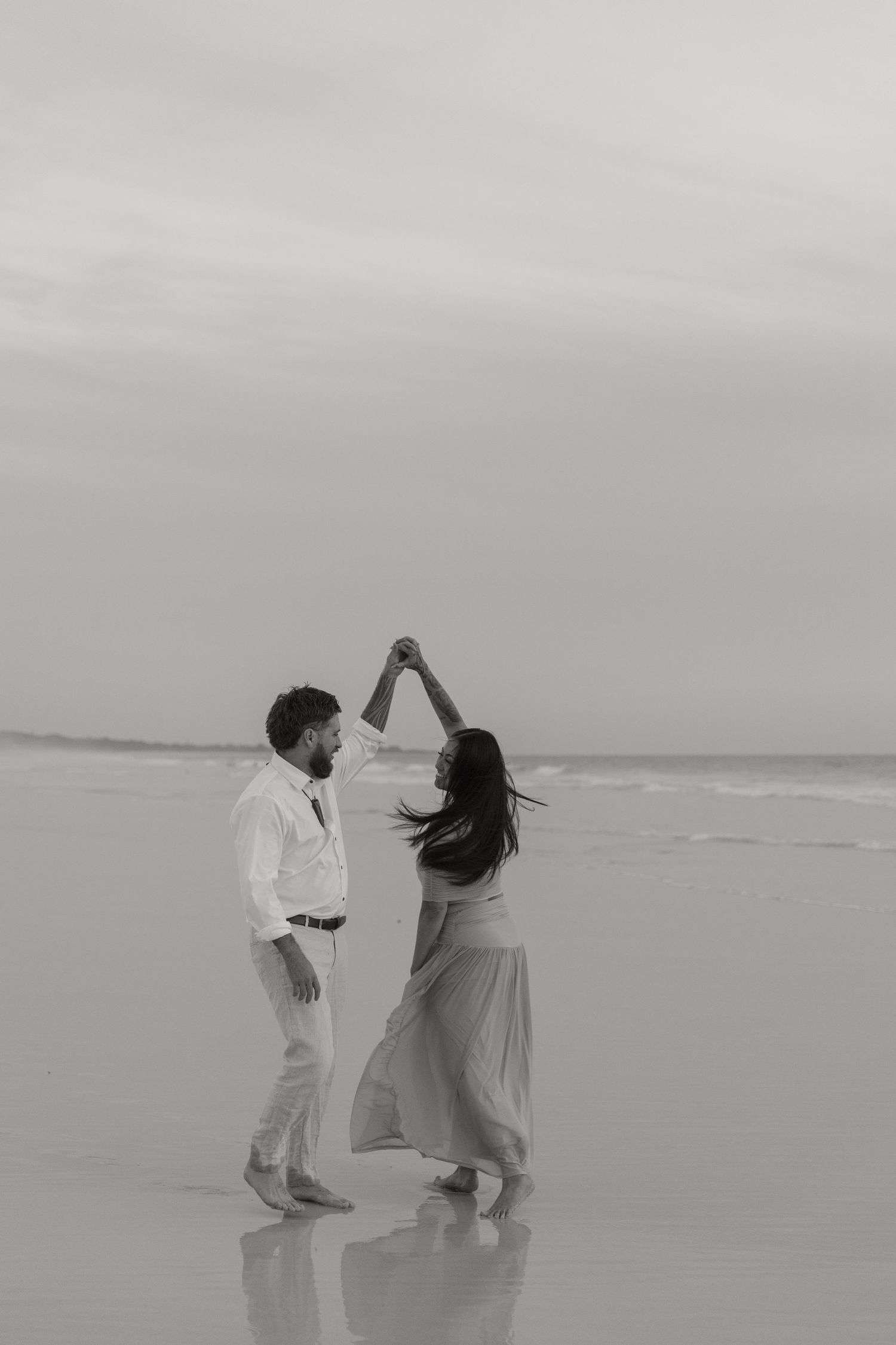 Pregnant woman in white maxi dress cradling baby bump on sandy beach at sunset.