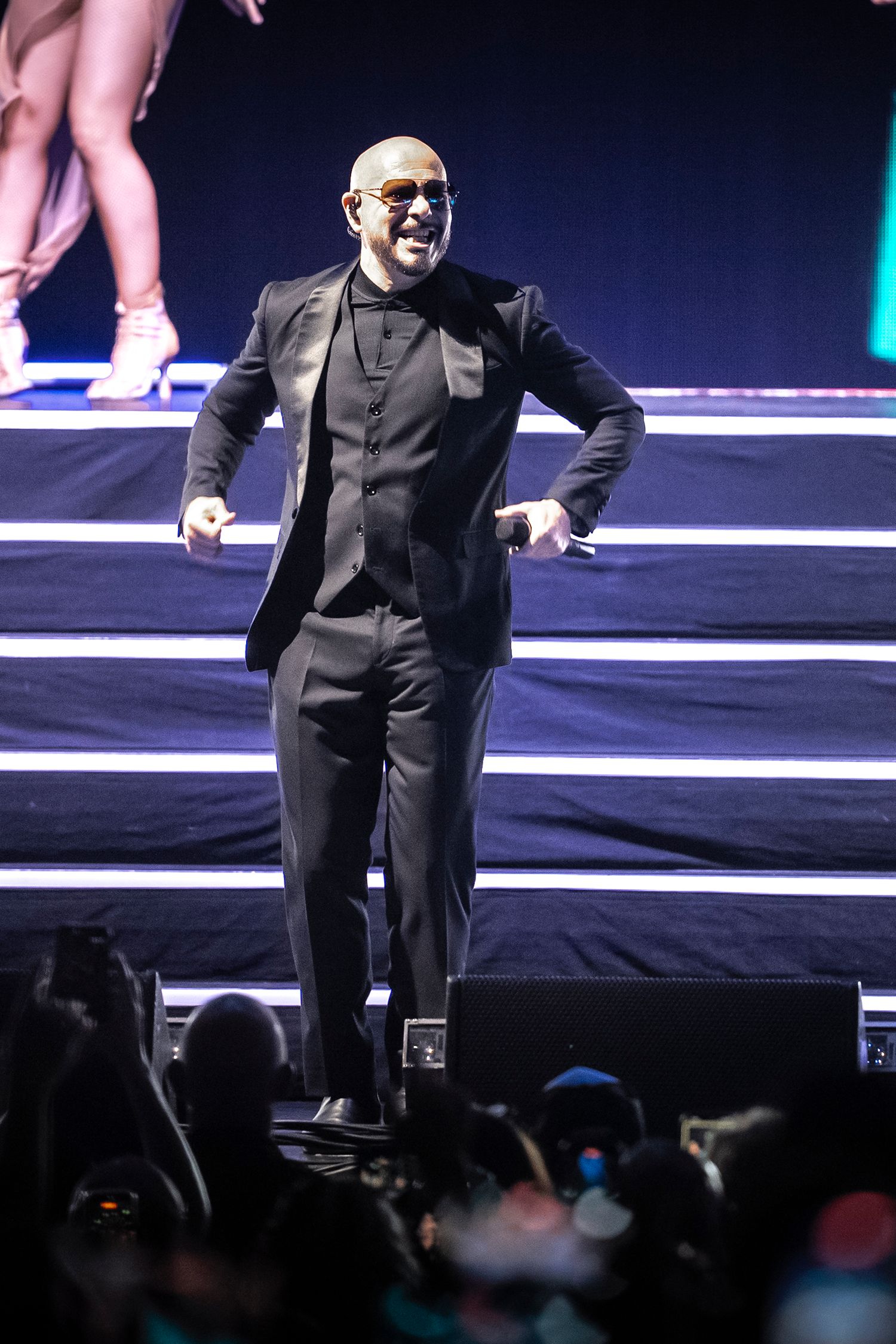 Bald performer in black suit and sunglasses on stage with dramatic lighting and energetic pose.
