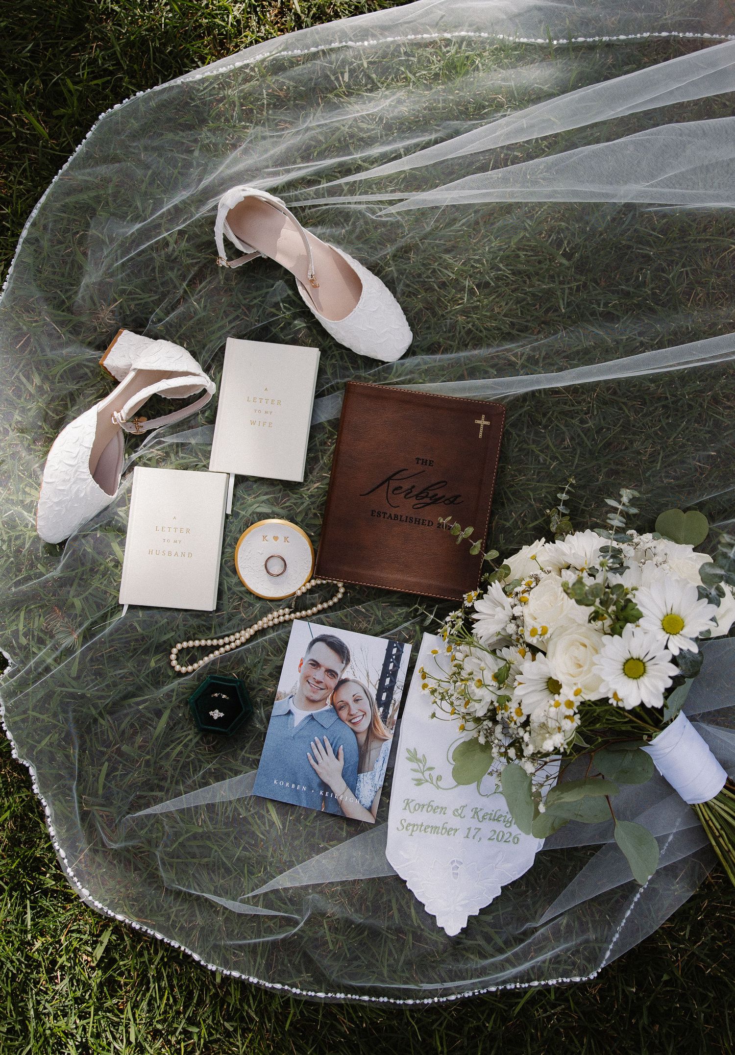 Wedding flat lay with veil, shoes, bouquet, rings, and personalized items on grass.