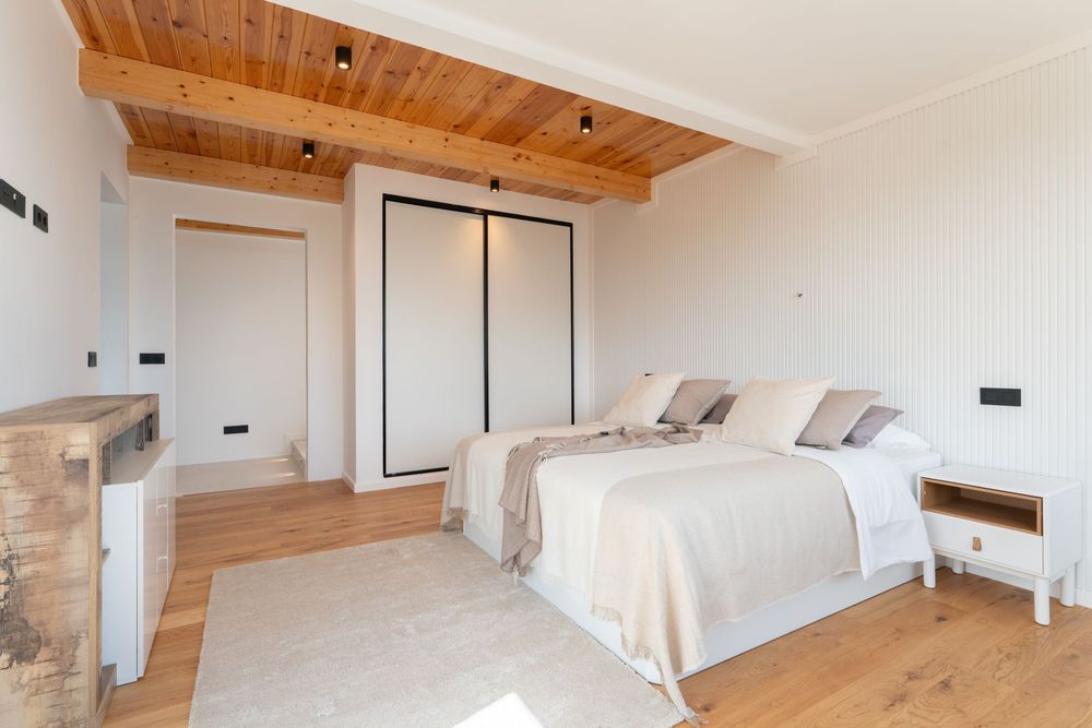 Bright bedroom with wooden ceiling beams, light hardwood floors, white walls, and a neutral-toned bed with elegant decor.