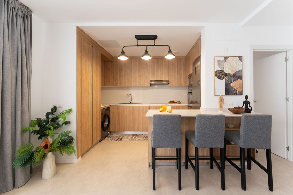 Modern open-plan kitchen and dining area with wooden cabinetry, gray bar stools, pendant lights, and tropical plant decor.