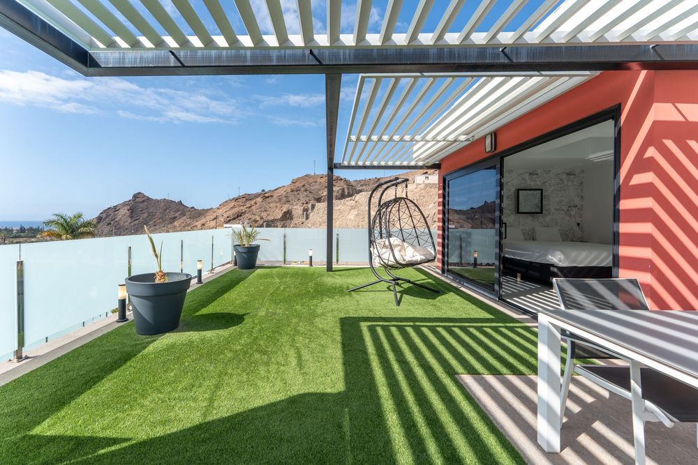 Modern rooftop terrace with artificial grass, pergola, hanging chair, and mountain views under clear blue sky.