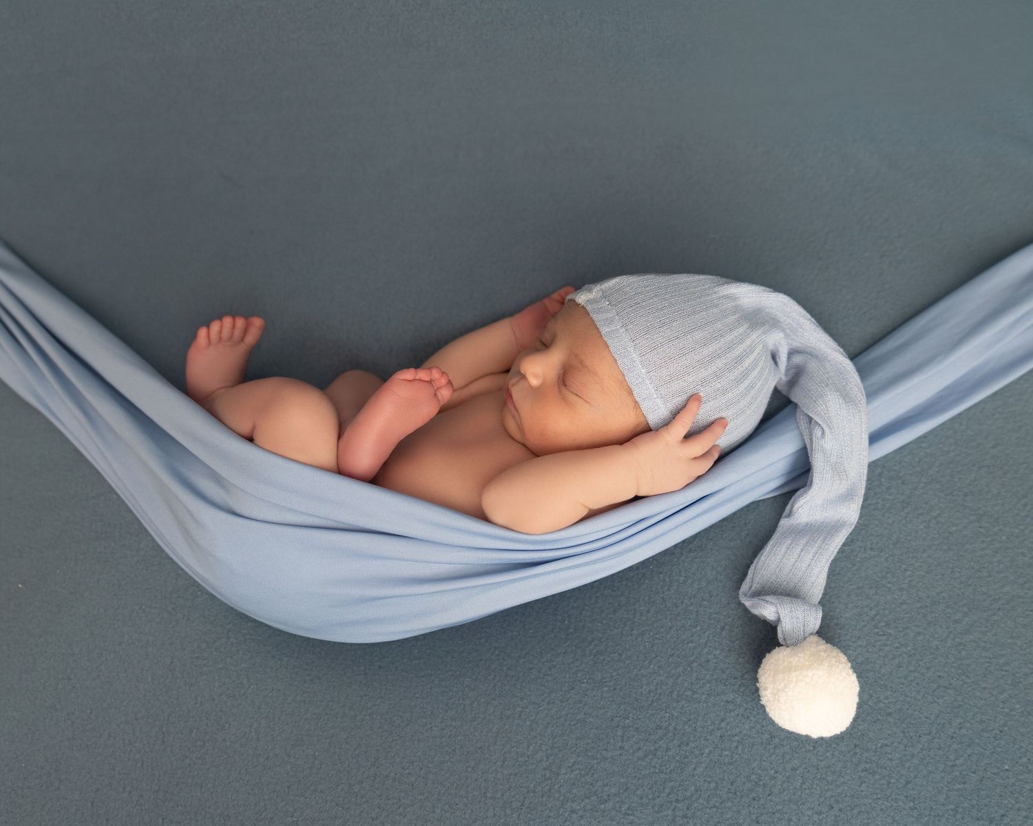 Newborn baby sleeping peacefully in a blue fabric hammock wearing a knit nightcap with pompom.