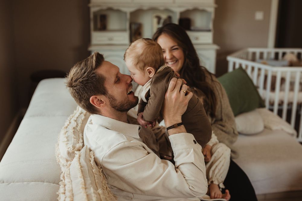 Happy young family with baby sitting on bed, parents smiling and playing with toddler in cozy bedroom.
