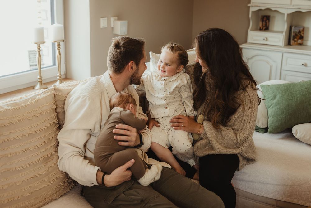 Family of four sitting together on a cozy sofa, parents smiling at toddler girl while holding a baby.