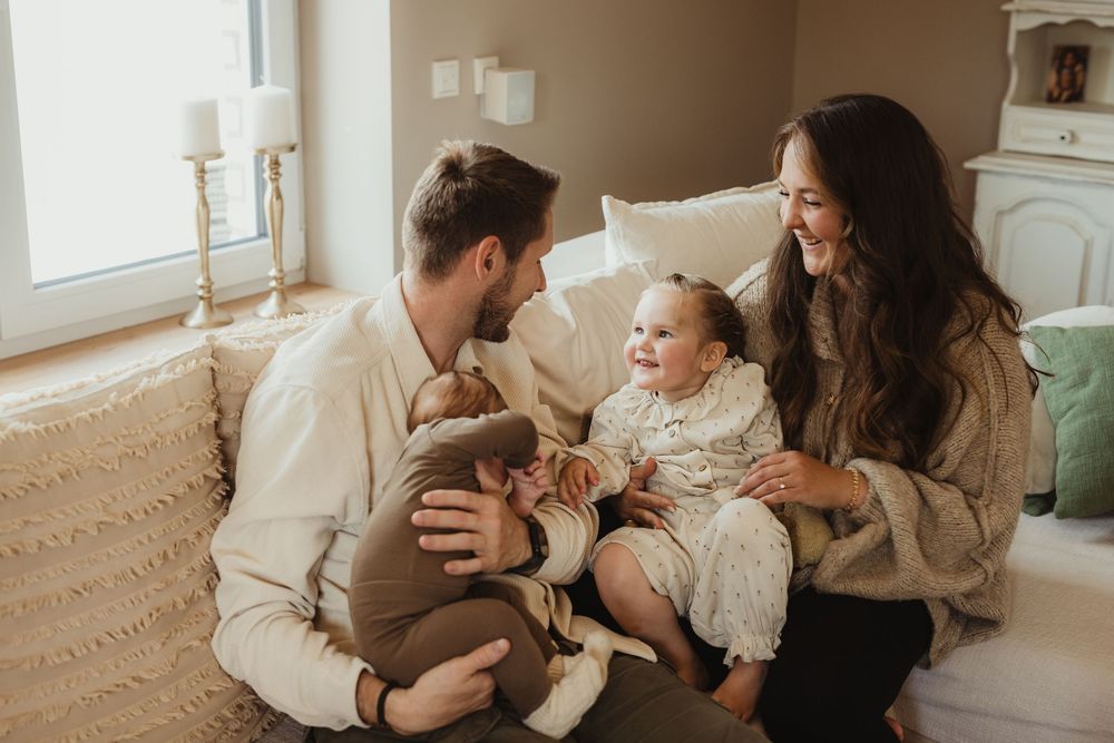 Happy young family with two babies sitting on a couch in a bright cozy living room, smiling and bonding together.