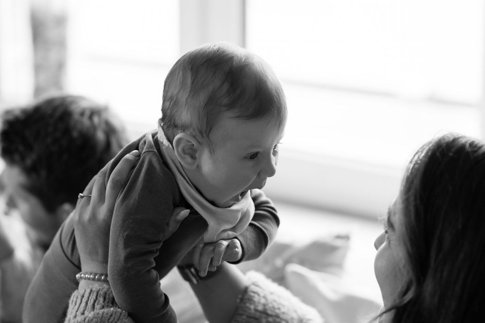 Black and white photo of a mother lifting her smiling baby in the air during a tender indoor moment.