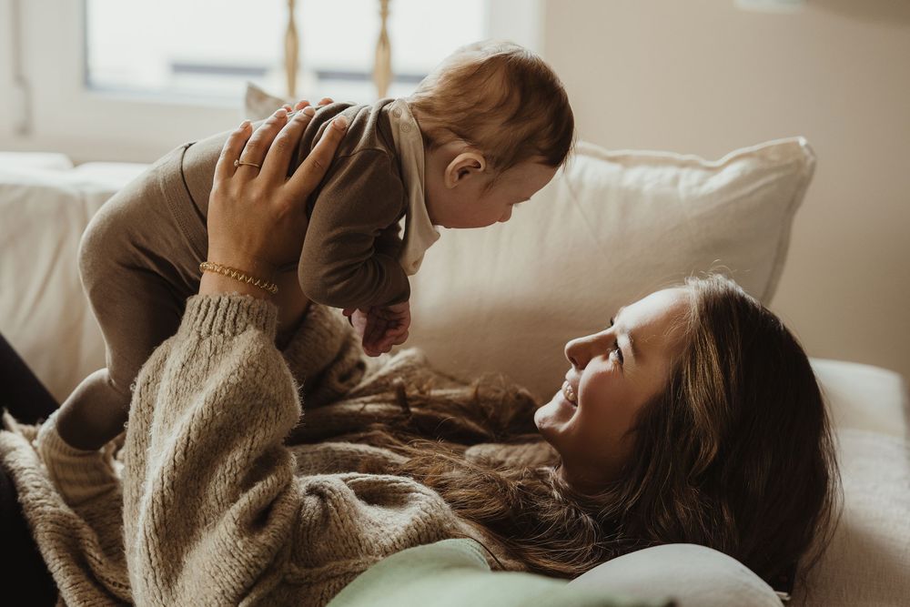 A mother lies on a couch smiling up at her baby she holds above her in a warm, cozy living room.