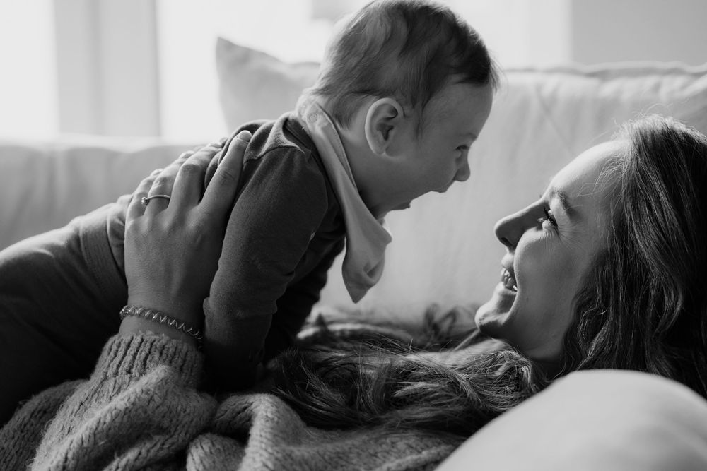 Black and white photo of a smiling mother lying down, lovingly holding her baby up face-to-face on a cozy sofa.