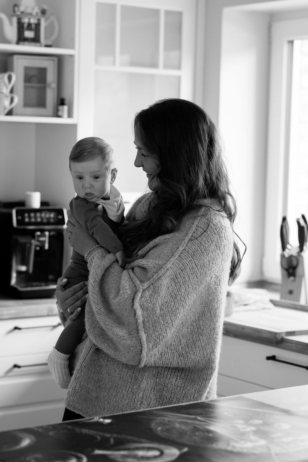 Black and white photo of a smiling mother holding her baby in a bright modern kitchen.