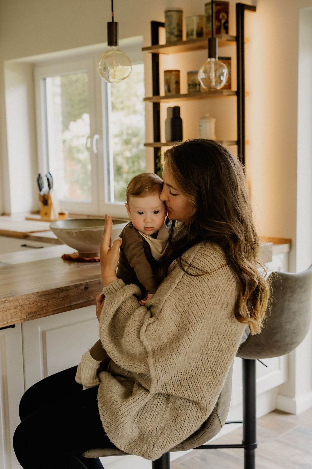 A mother in a beige knit sweater sits in a bright kitchen holding her baby on her lap.