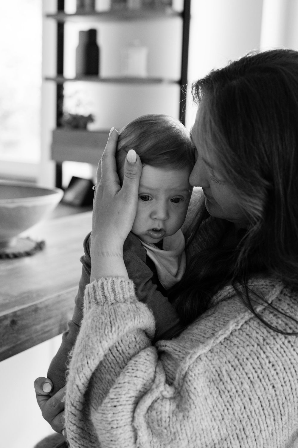 Black and white photo of a mother tenderly holding and kissing her baby in a cozy home interior.