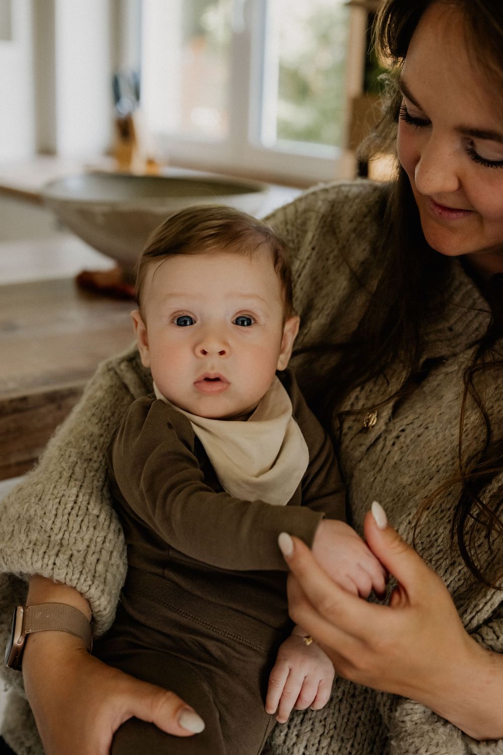 A smiling mother holds her wide-eyed baby dressed in olive green, sharing a tender moment indoors.