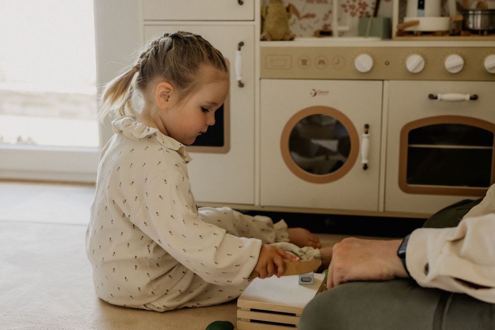 Young girl sitting on floor playing with toys near a white wooden play kitchen in a bright room.