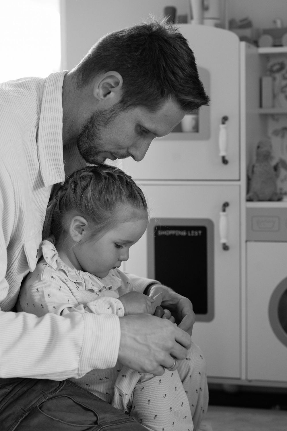 Black and white photo of a father sitting with his young daughter in a playroom, both looking down tenderly.