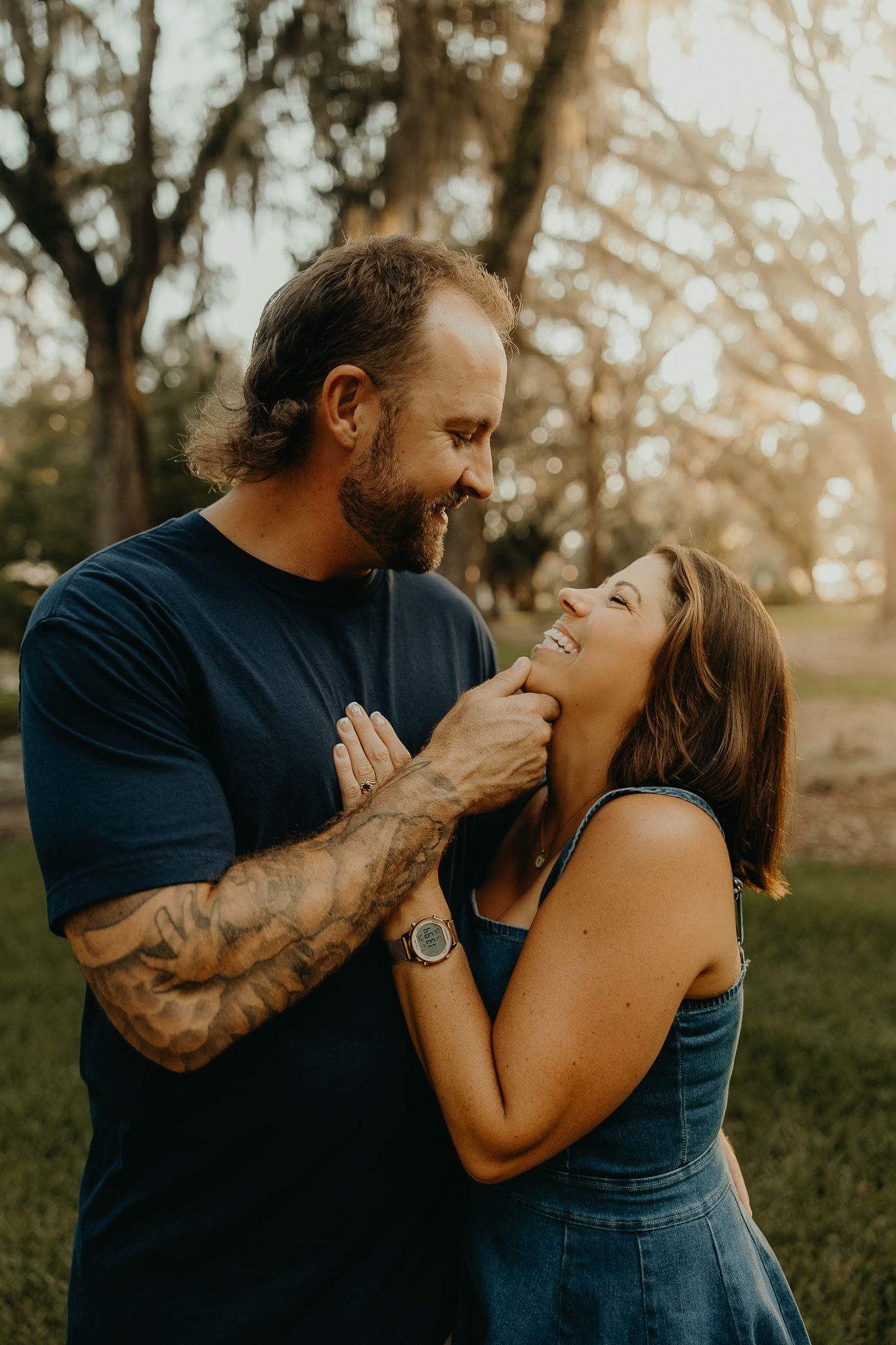 Couple laughing and holding hands outdoors at golden hour, man with tattoos in navy shirt, woman in denim overalls.