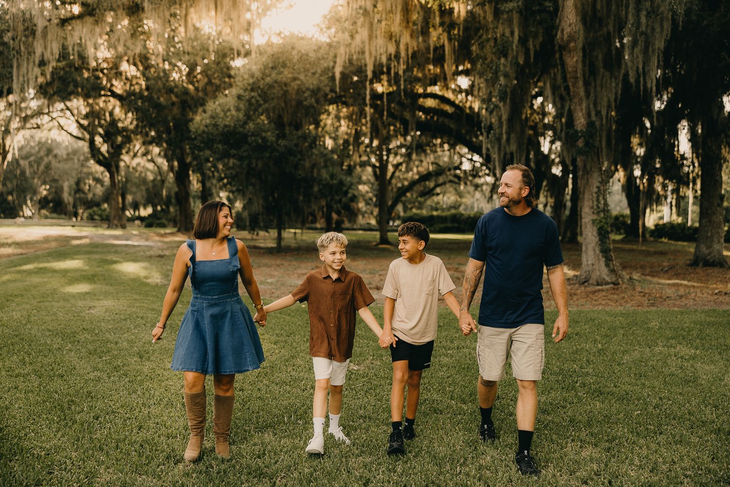 Family of four holding hands walking through a lush green park with Spanish moss-draped oak trees at golden hour.
