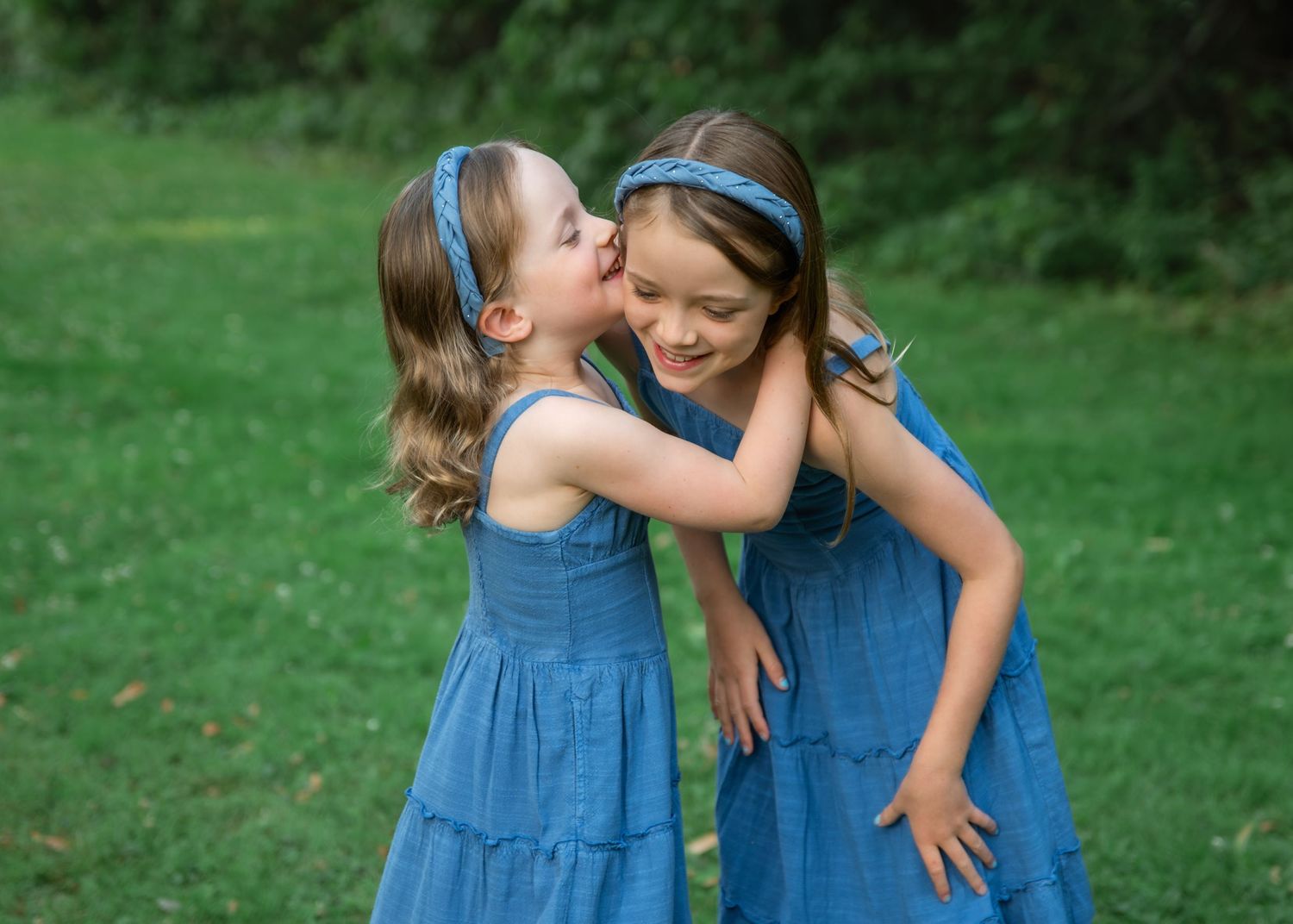 Two young girls in matching blue dresses and headbands share a sweet moment, one whispering to the other outdoors.