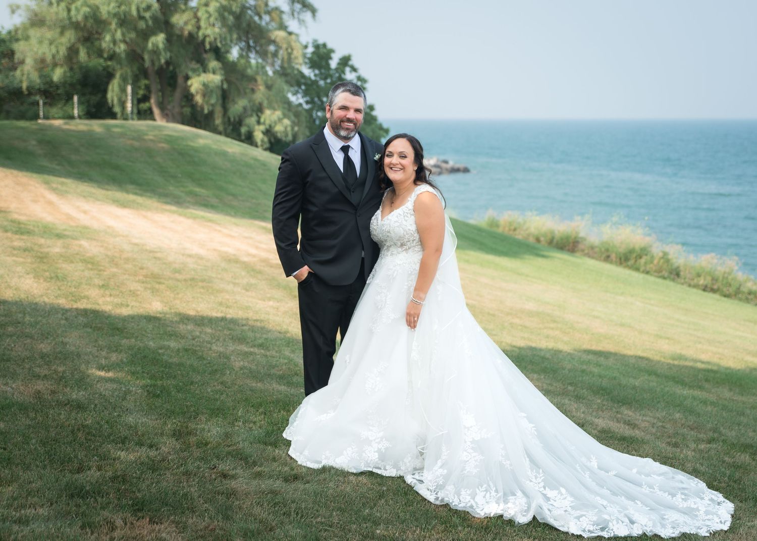 Bride in white gown and groom in black tuxedo pose on green hillside with ocean view on wedding day.