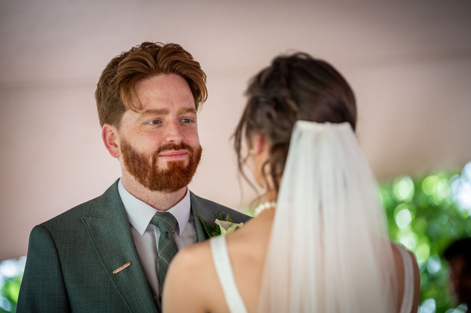 Groom in green suit smiling at bride in white veil during outdoor wedding ceremony.