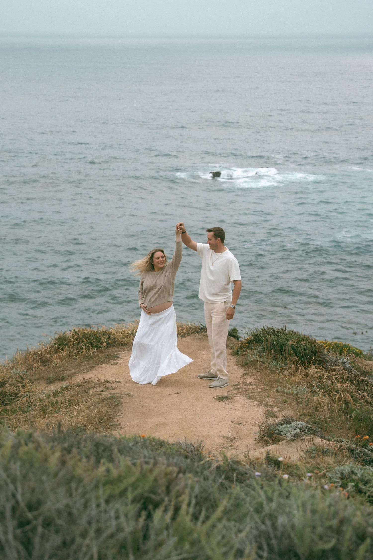 Couple dancing on a coastal cliff overlooking the ocean, wearing neutral tones, with waves and green shrubs in the background.