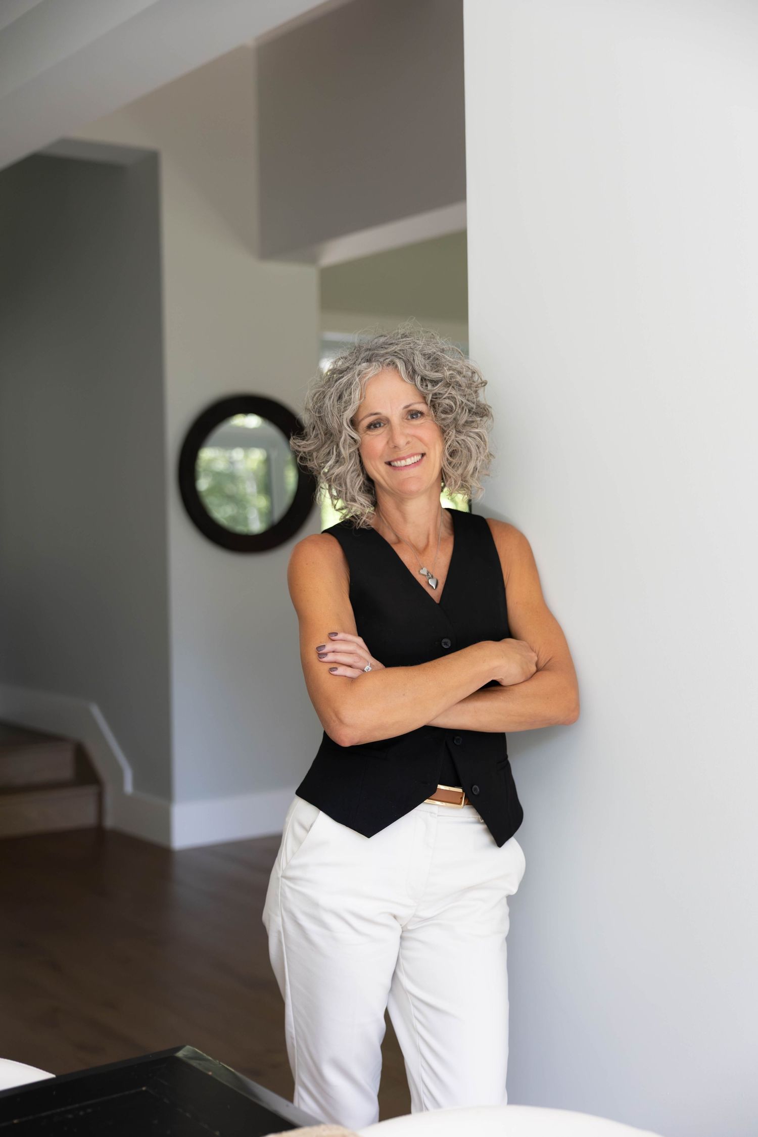 Smiling middle-aged woman with gray curly hair, arms crossed, wearing black top and white pants, leaning against wall indoors.