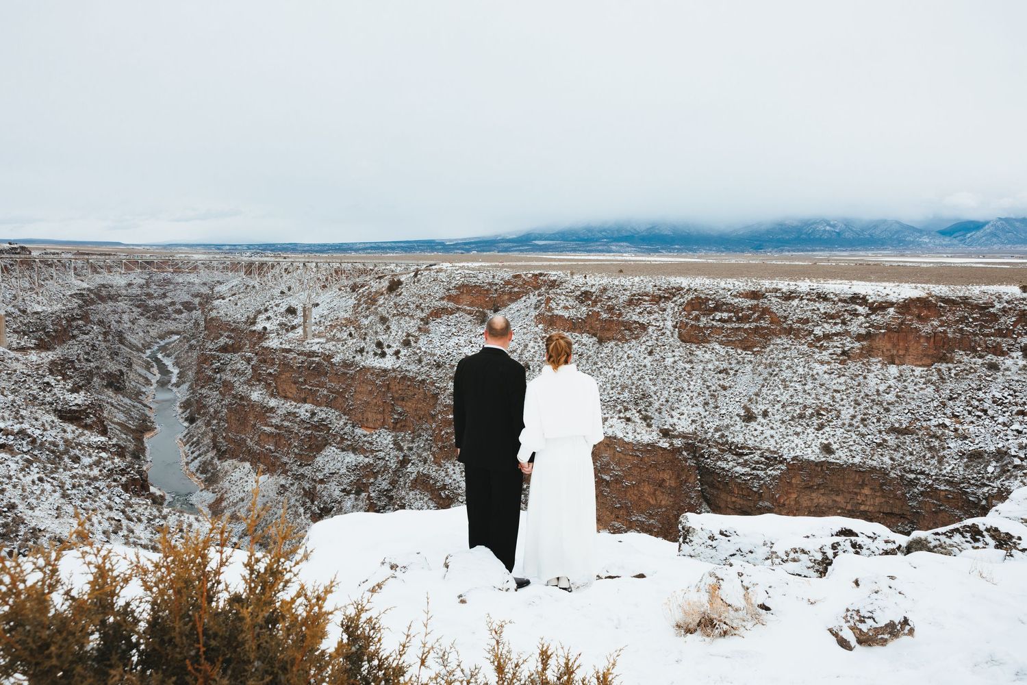 Couple embraces at snowy Taos Gorge overlook in New Mexico, bride in white coat, groom in black suit.