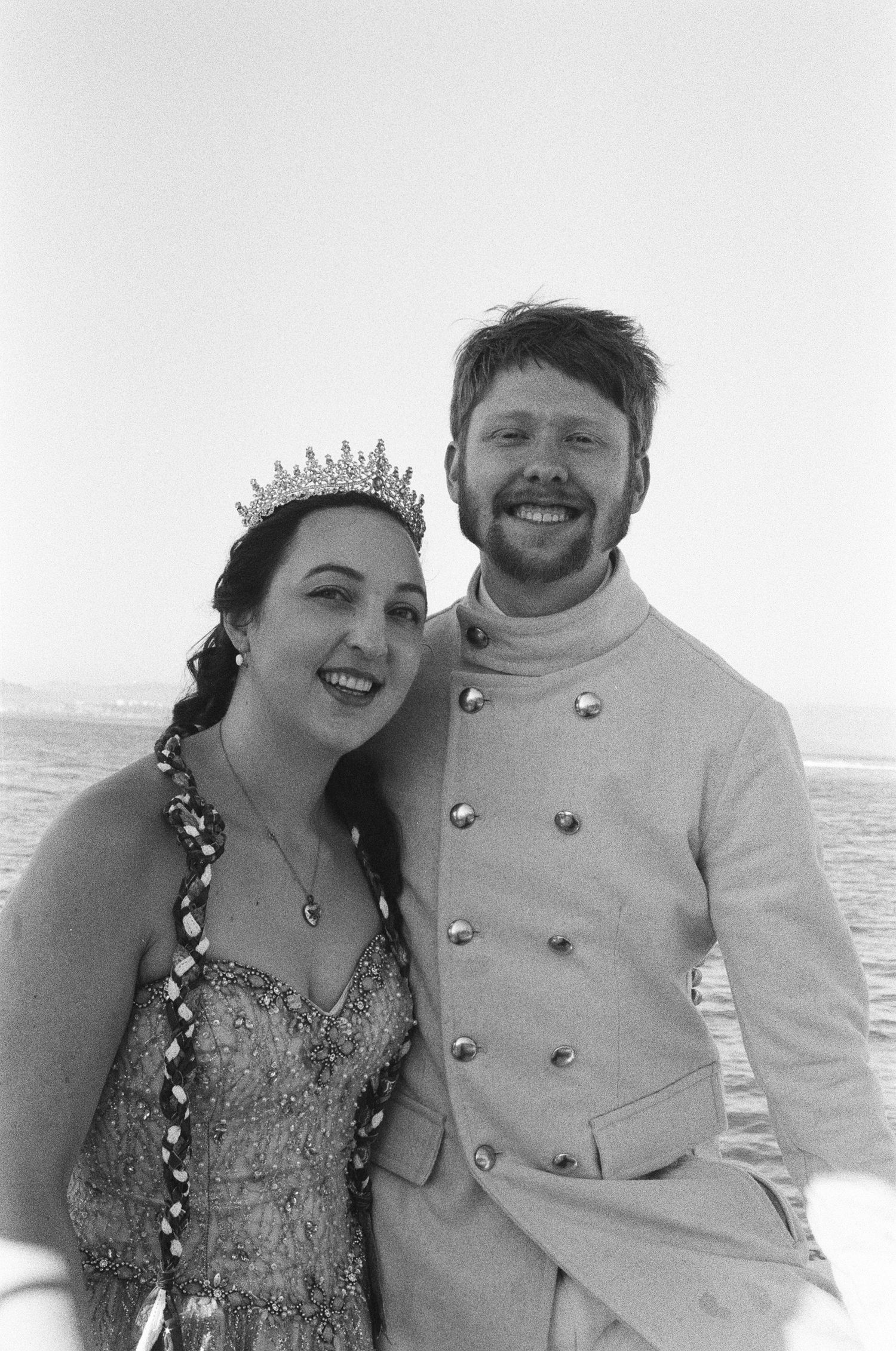 Black and white photo of a smiling couple, woman wearing tiara and beaded gown, man in military-style jacket, outdoors.
