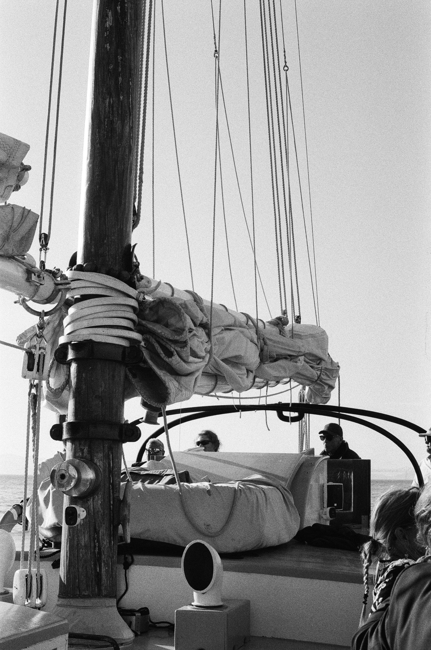 Black and white photo of sailors on a classic sailboat deck with rigging, ropes, and furled sails against a calm sea horizon.