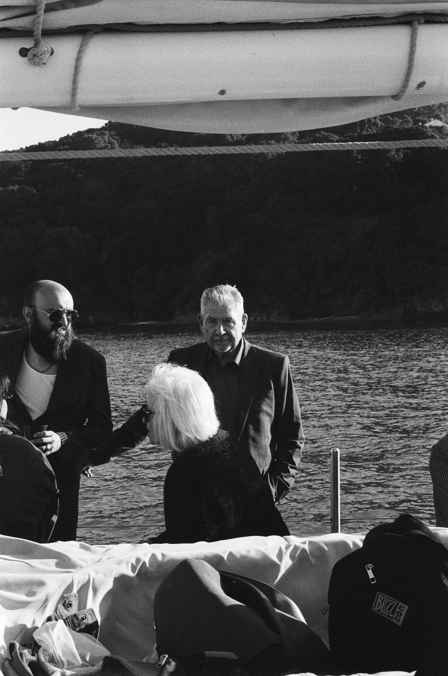 Black and white photo of three people conversing on a boat deck near water, with a lifeboat visible above.
