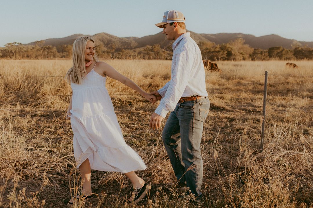 Couple holding hands walking in dry golden field at sunset with mountain views.