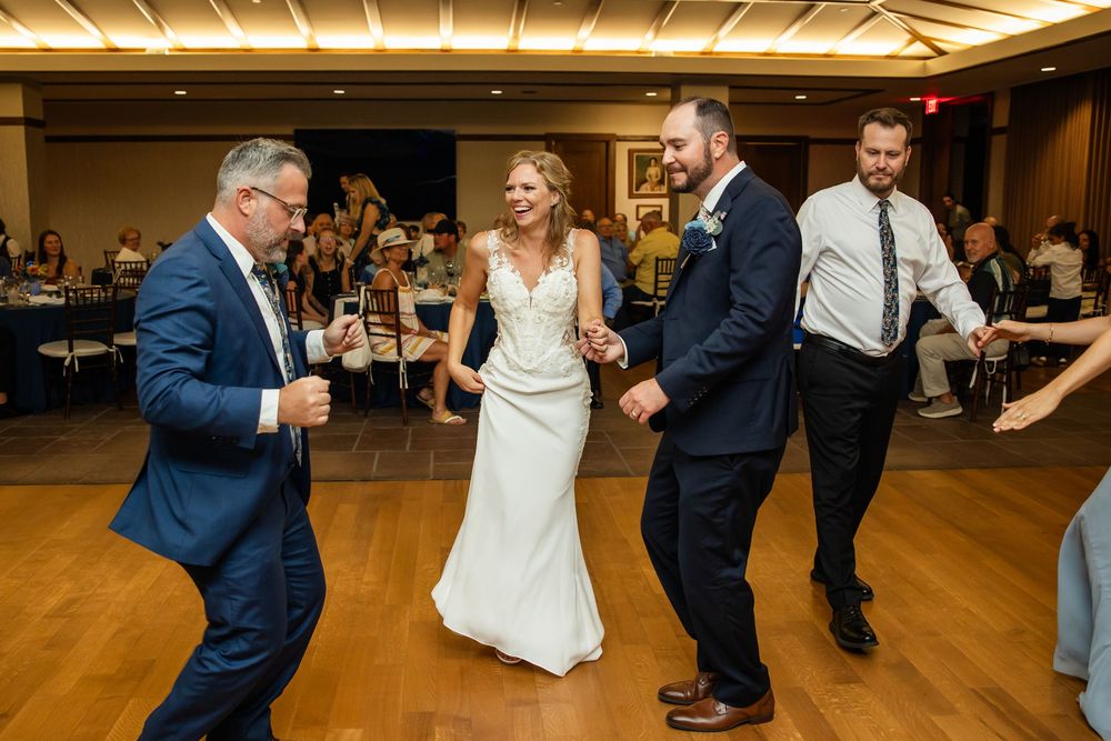 Bride in white gown dances joyfully with groom and groomsmen in navy suits at elegant wedding reception.
