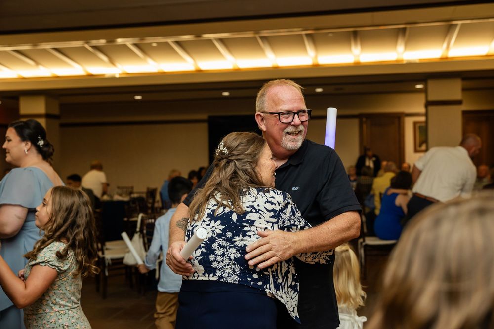 Man in black shirt hugging woman in floral dress at a wedding reception celebration.