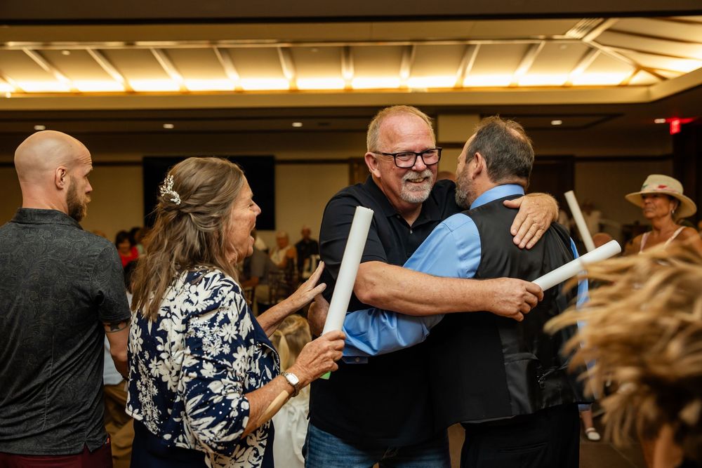 People hugging and celebrating at an indoor event, smiling man in blue shirt embracing another guest in a crowded venue.
