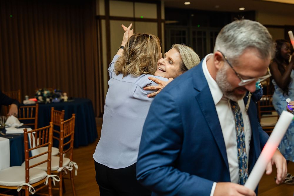 Two women embrace warmly at an elegant event venue while a man in a blue suit stands nearby holding papers.