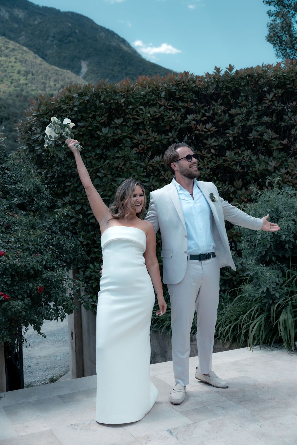 Joyful bride in white strapless gown raises bouquet as groom in tan suit celebrates outdoors with mountain backdrop.