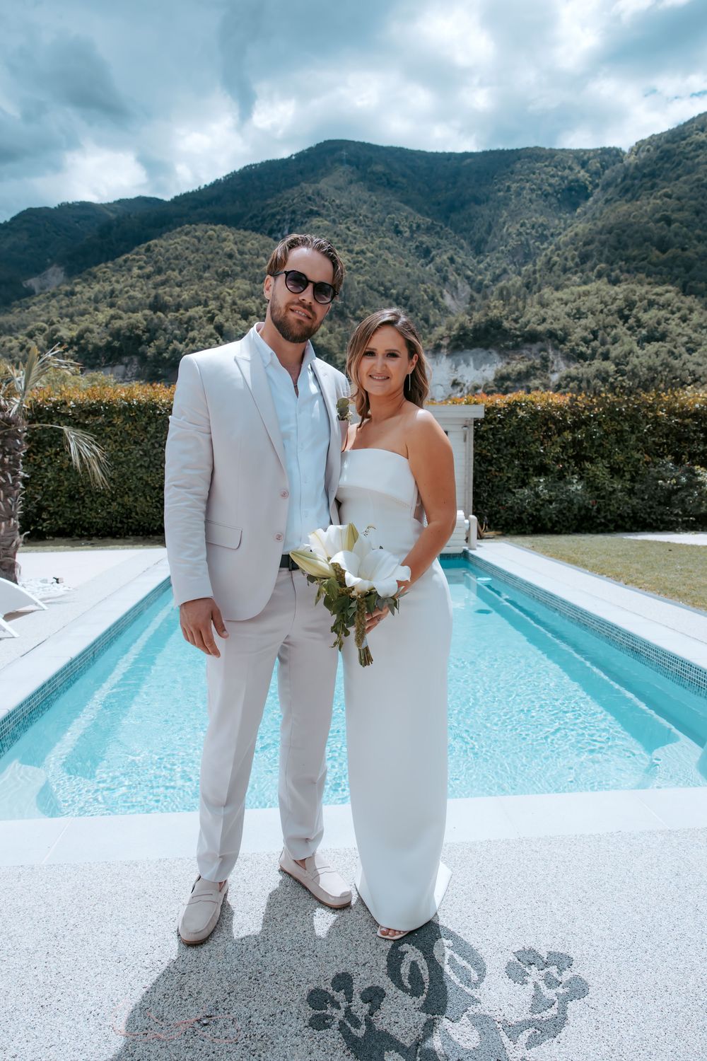 Bride and groom in white attire pose by a pool with mountain backdrop on their wedding day.