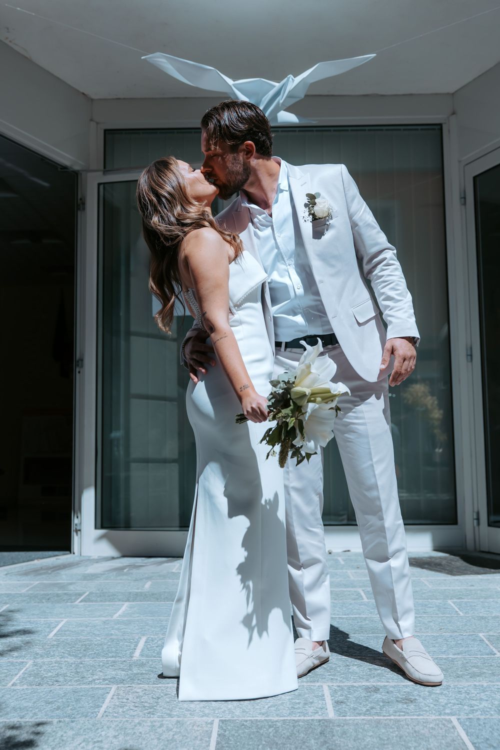 A couple shares a romantic kiss in wedding attire, bride in white gown holding bouquet, groom in light gray suit.