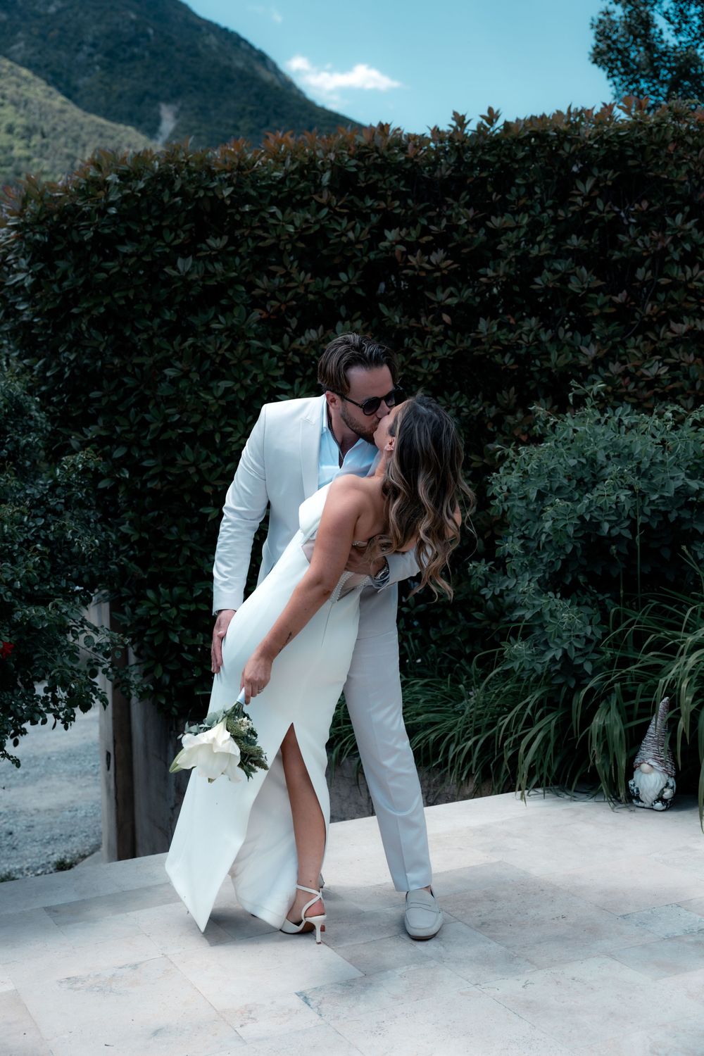 A bride and groom share a romantic dip kiss outdoors, dressed in white wedding attire with lush green hedges behind them.