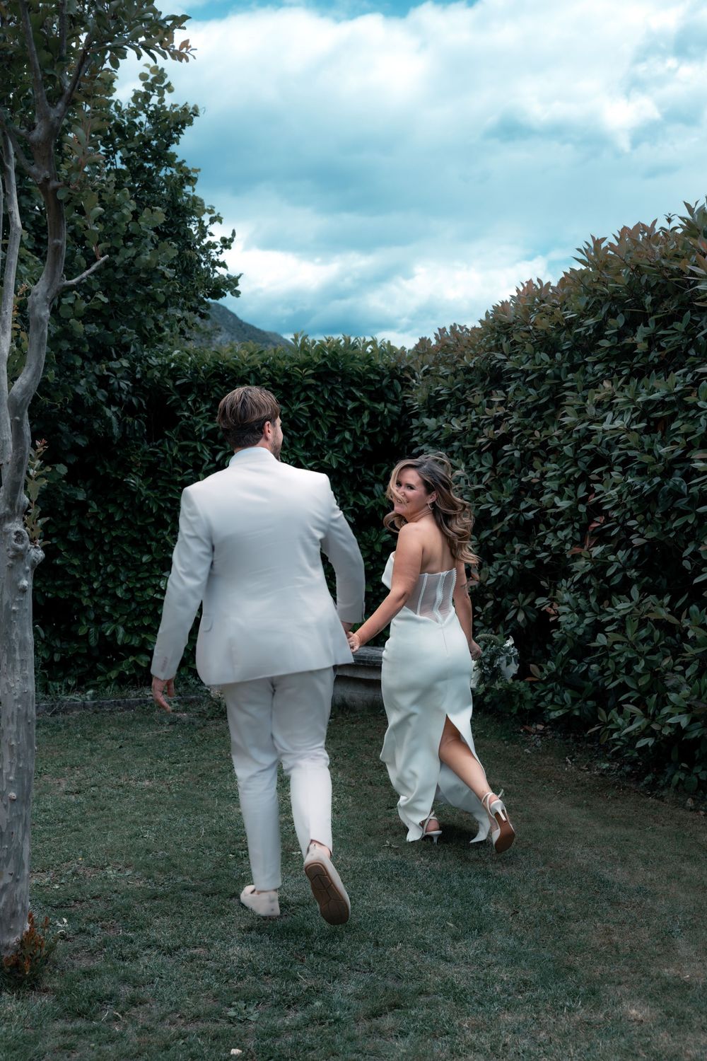 Bride and groom walking hand in hand through lush garden greenery, bride in white dress glancing back, groom in white suit.