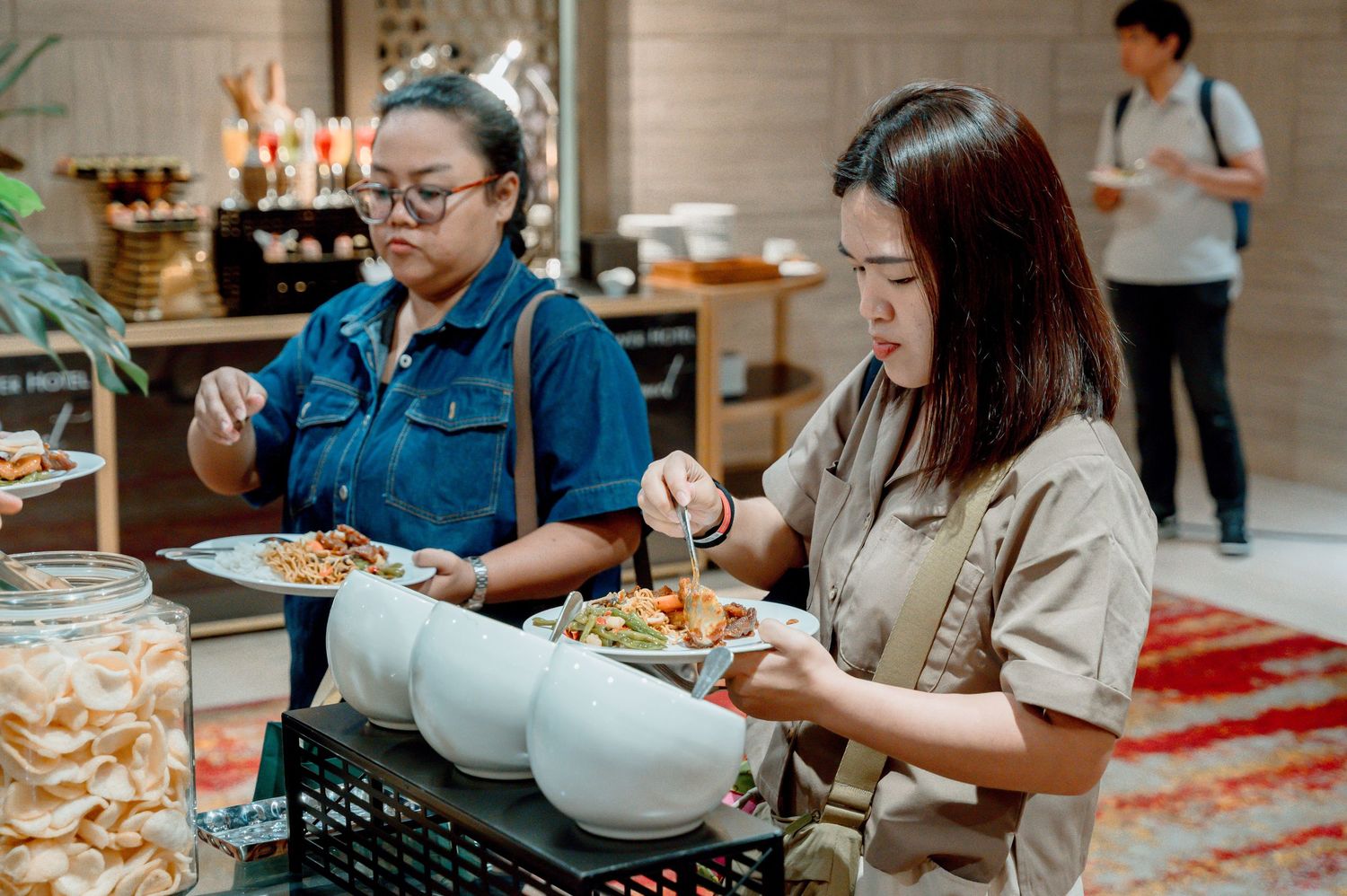 Two women serving themselves at a buffet, selecting food from large serving bowls in a restaurant setting.