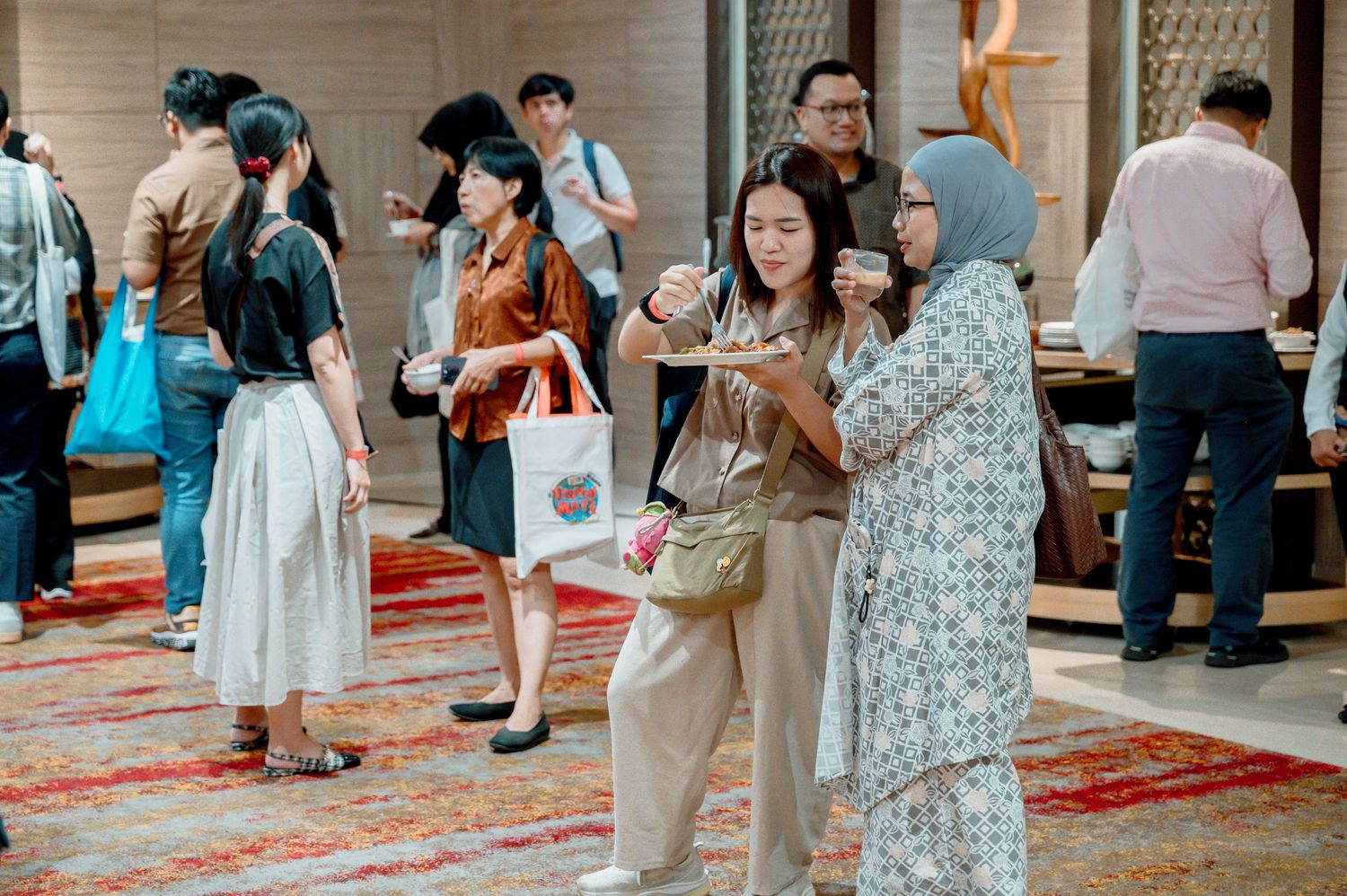 Diverse group of people networking at an indoor event, with two women in foreground sharing food and conversation.