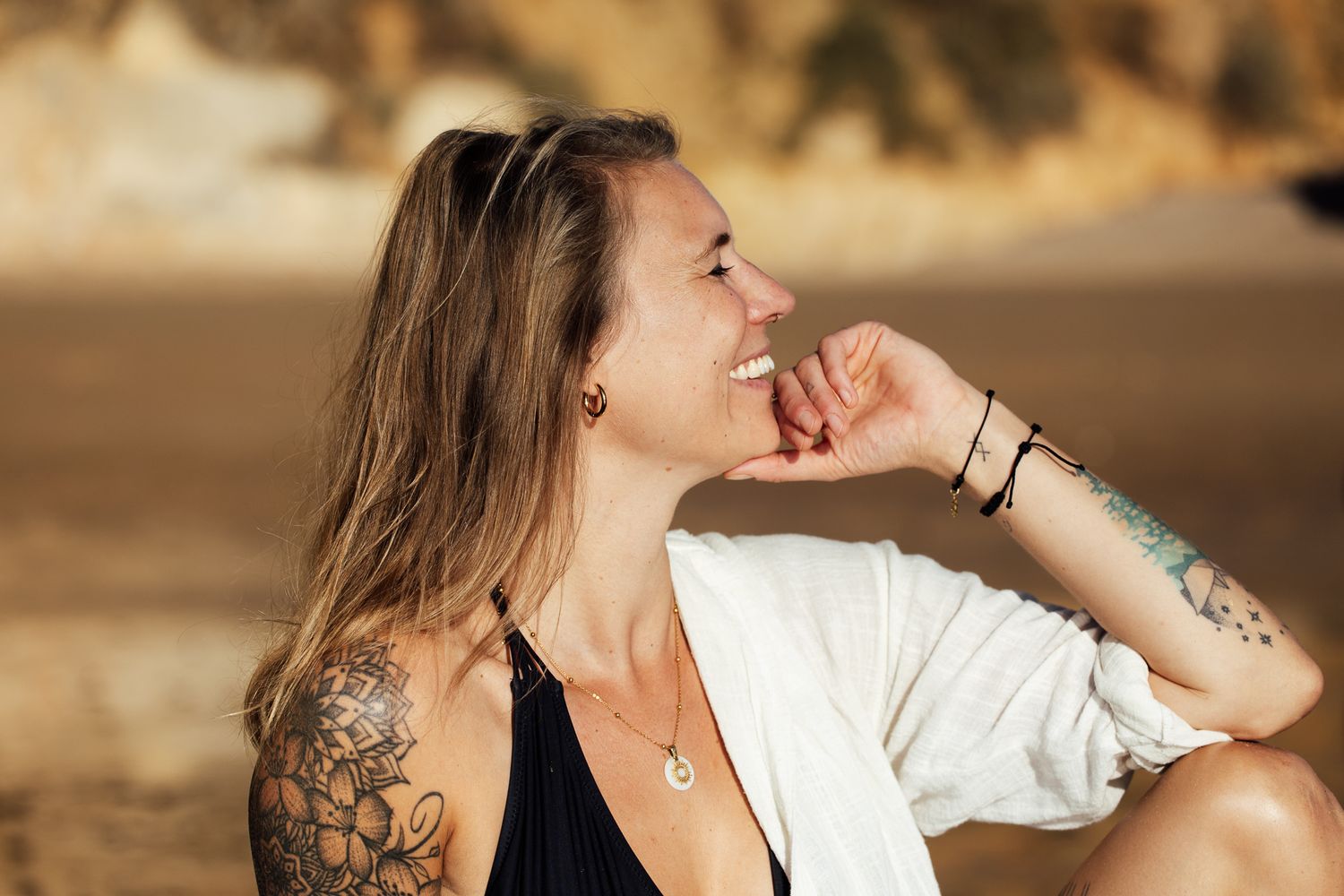Smiling woman with tattoos sitting on a beach, wearing a white cardigan and black top, enjoying a sunny day.