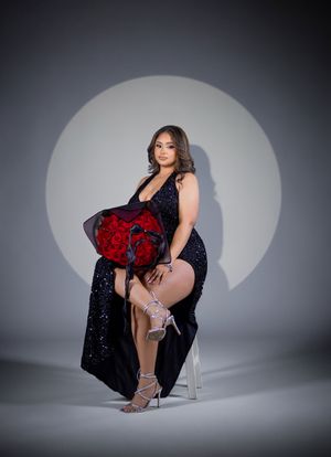 Woman in black sequin gown holding red roses bouquet, seated elegantly against spotlight background.