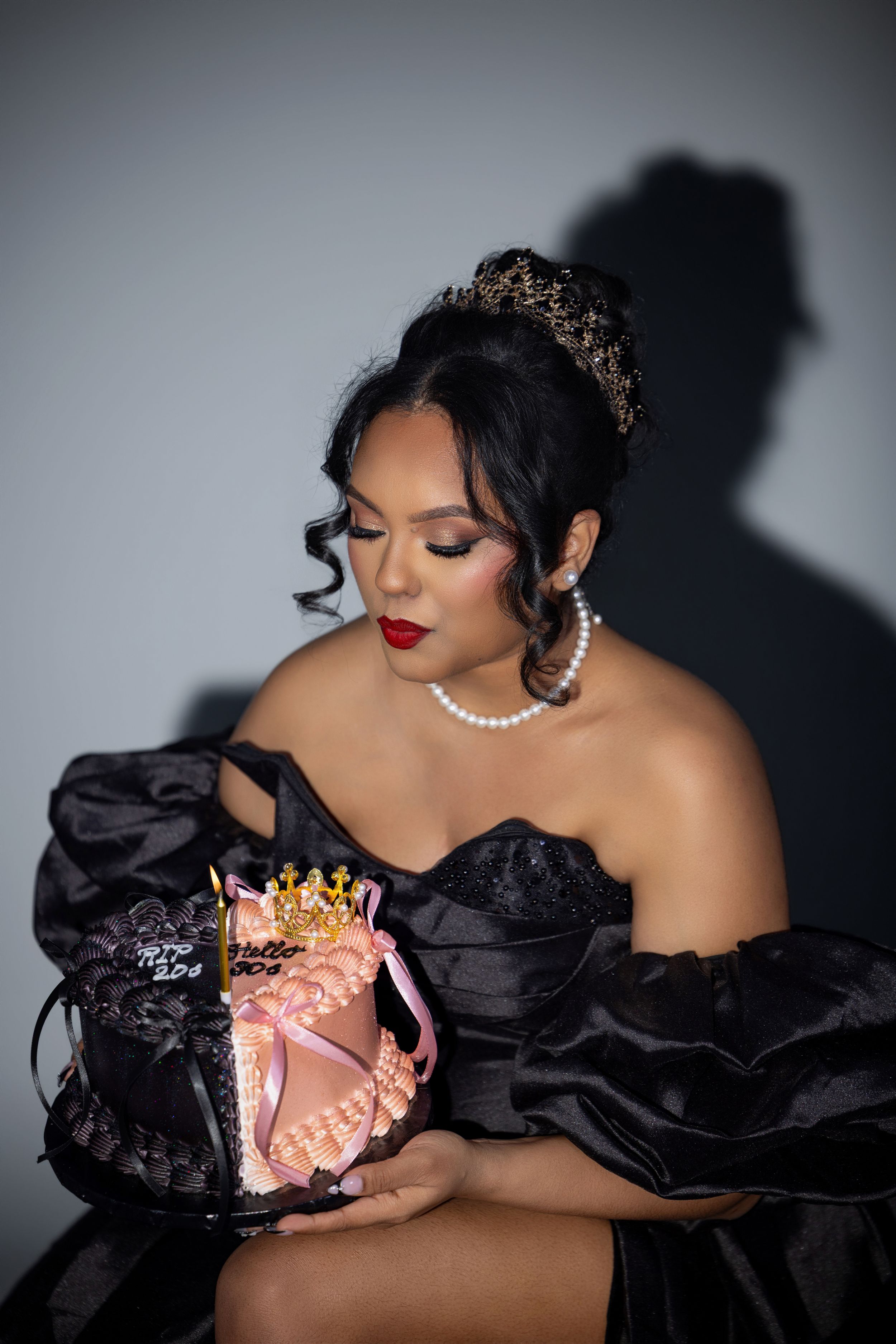 Woman in black off-shoulder gown with tiara holds decorative pink cake, wearing pearl jewelry and red lipstick.